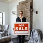 Female real estate agent indoors holding a 'For Sale' sign, showcasing property listing.